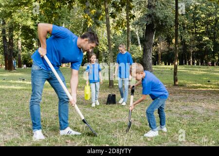 Vater und Sohn graben Boden, während Mutter und Tochter tragen jungen Baum und Gießkanne, Ökologie Konzept Stockfoto