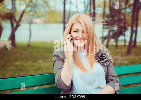 Portrait junge schöne Frau sprechen auf Handy sitzen auf einer Bank in einem Stadtpark grünen Bäumen Hintergrund. Gesicht mit toothy Lächeln. Wunderschönes Mittelfeld Stockfoto