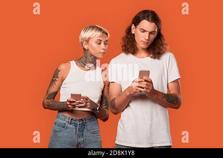Freistil. Junge tätowierte Paar stehend isoliert auf orange Freundin Blick auf Freund Smartphone neugierig Stockfoto
