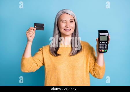 Foto von positiven alten Frau halten Kreditkarte Maschine Leser Empfehlen Bank Kauf Wink Blink tragen Stil stilvolle trendige Pullover Isoliert über blau Stockfoto