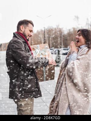 Mann, der seiner Freundin ein Weihnachtsgeschenk gibt. Emotional überrascht glückliches Mädchen erhält ein Geschenk draußen in schneebedeckten Wetter. Stockfoto