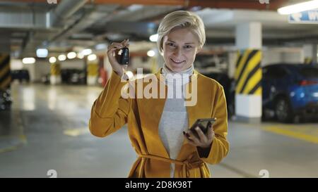 Junge blonde modische Frau mit Autoschlüssel und Smartphone in der Tiefgarage. Hochwertige Fotos Stockfoto