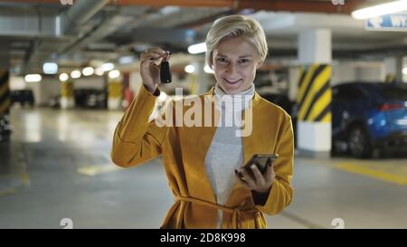 Junge blonde modische Frau mit Autoschlüssel und Smartphone in der Tiefgarage. Hochwertige Fotos Stockfoto
