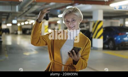 Junge blonde modische Frau mit Autoschlüssel und Smartphone in der Tiefgarage. Hochwertige Fotos Stockfoto
