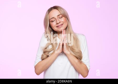 Studio Portrait von charmanten blonde Frau schließt die Augen und hält die Handfläche zusammen, hofft auf das Beste im Leben, betet um Glück zu haben, um Hilfe zu bitten Stockfoto