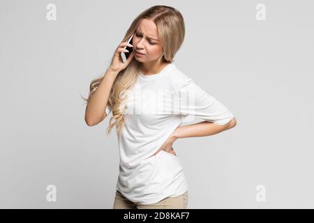 Kaukasische Frau, die Schmerzen hat, sprechen auf dem Handy Muskel oder chronische Nervenschmerzen in ihrem Rücken nach der Arbeit, isoliert. Erkrankungen der Wirbelsäule, Skoliose, Osteo Stockfoto