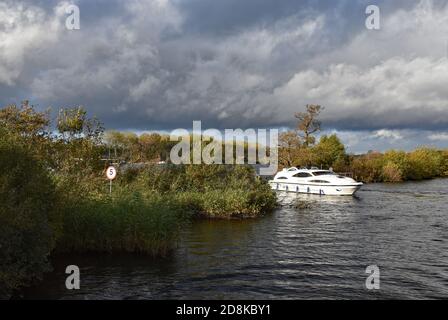 Ein kleines Boot taucht von Wroxham Broad auf, um den Fluss Bure zu verbinden, während sich Sturmwolken über dem Wasser sammeln. Ein 5 Meile eine Stunde Zeichen ist in den Büschen, England gesehen. Stockfoto