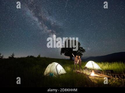 Mann Wanderer mit Freundin zeigt auf Milchstraße auf majestätischen Sternenhimmel. Liebespaar steht in der Nähe von beleuchteten Zeltzelten und blickt auf den Himmel mit Sternen. Konzept des Wanderns, Nachtcamping, Beziehungen Stockfoto