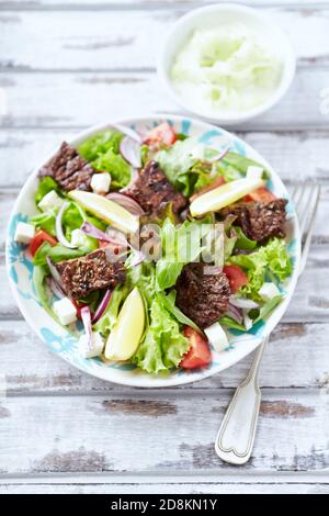 Schmackhafter Salat mit gegrillten Rinderstücken, Kirschtomaten, Feta-Käse und Kalamata-Oliven. Heller Holzhintergrund. Stockfoto