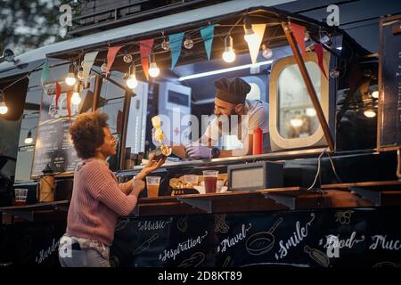 Männliche Angestellte im Fast-Food-Service lachend, gesellig mit afro-amerikanischen weiblichen Kunden, während sie Senf in ihr Sandwich Stockfoto