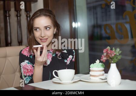 Porträt eines Mädchens, das die Spracherkennung des Mobiltelefons nutzt und in der Nähe eines Fensters eines Restaurants oder eines Cafés aufschaut. Menschen und Technologie Stockfoto