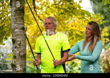 Ein Mann in den späten Sechzigern trainiert im Herbst mit einer Widerstandsband mit Hilfe einer jüngeren weiblichen Personal Trainerin im Freien. Stockfoto