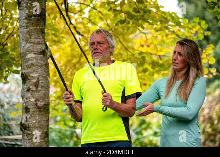 Ein Mann in den späten Sechzigern trainiert im Herbst mit einer Widerstandsband mit Hilfe einer jüngeren weiblichen Personal Trainerin im Freien. Stockfoto