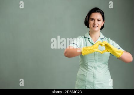 Junge Putzfrau trägt ein grünes Hemd und gelbe Handschuhe, tun Herzzeichen mit ihren Händen isoliert auf grünem Hintergrund Stockfoto