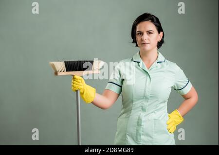 Junge Putzfrau trägt ein grünes Hemd und gelbe Handschuhe, hält Besen isoliert auf grünem Hintergrund Stockfoto