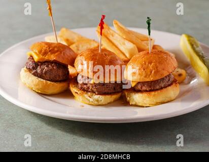 Drei Cheeseburger-Slider mit Pommes Frites und einer Gurke Eine weiße Platte Stockfoto