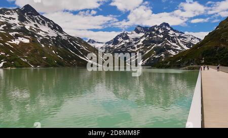 Touristen genießen die schöne Aussicht von Silvretta Stausee Damm, Montafon, Österreich mit schneebedeckten Bergen (einschließlich Biz Buin) im Hintergrund. Stockfoto