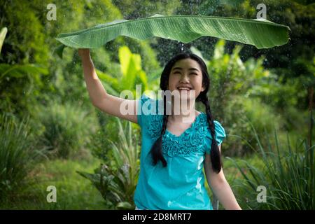 Porträt einer jungen Asienfrau mit schwarzem Haar, die ein Bananenblatt im Regen auf dem grünen Gartenhintergrund hält Stockfoto