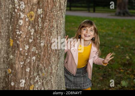 Zahnlose lustige niedliche Mädchen spielen verstecken und suchen im Herbst Park. Blickt hinter einen Baum mit einem verspielten Lächeln und lacht. Saisonales Konzept Stockfoto