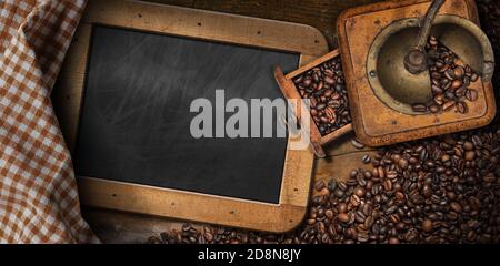 Alte manuelle Kaffeemühle aus Metall und Holz mit gerösteten Kaffeebohnen auf dem Tisch mit leerer Tafel und karierter Tischdecke. Stockfoto