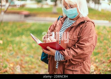 Erwachsene kaukasische blonde Frau in lässiger Kleidung und Schutzmaske Steht, die Notizen in Notizbuch mit Bleistift in einem Park Stockfoto