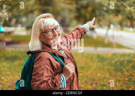Erwachsene blonde Frau in bequemer Kleidung mit Rucksack zeigt die Richtung im Park mit dem Finger. Touristenreisender in einem wunderschönen Ort Stockfoto