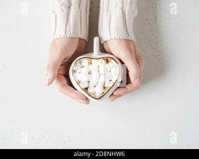 Weibliche Hände halten weiße Keramik-Tasse in Form von Herz mit heißem Getränk und Marshmallows auf hellem Hintergrund. Draufsicht. Speicherplatz kopieren Stockfoto