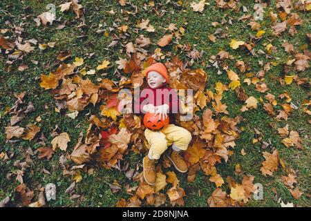 Das Mädchen liegt auf dem Rücken auf den gelben Herbstblättern mit einem großen Kürbis zwischen den Beinen und lacht fröhlich. Stockfoto