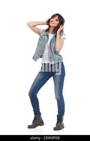 Junge Frau in Jeans Unknopfed Weste steht und Musik in Kopfhörer hören. Studioaufnahme in voller Länge, isoliert auf Weiß. Stockfoto