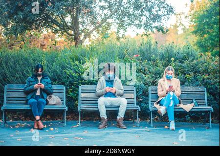 Gruppe von multirassischen Freunden mit Gesichtsmaske in der sozialen Distanzierung sitzen auf der Bank im Park in covid-19 Zeit - Stadt Ausbruch Lebensstil, schützendes Gesicht Stockfoto