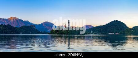 Ein Panoramabild des Bleder Sees, zentriert auf der Insel Bled, bei Sonnenuntergang, aufgenommen vom Rand. Stockfoto