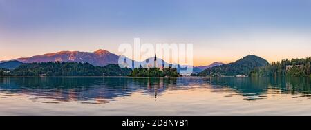 Ein Panoramabild des Bleder Sees, zentriert auf der Insel Bled, bei Sonnenuntergang, aufgenommen vom Rand. Stockfoto