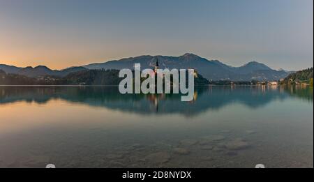 Ein Panoramabild des Bleder Sees, zentriert auf der Insel Bled, bei Sonnenuntergang, aufgenommen vom Rand. Stockfoto