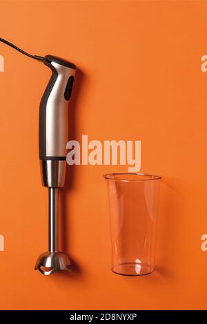 Stabmixer und leere Tasse auf einem karrot-orangefarbenen Hintergrund. Moderne elektrische Küchengeräte für die Herstellung von Saucen, Smoothies, Püree. Speicherplatz kopieren Stockfoto