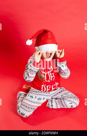 Ein glückliches Mädchen in roten Weihnachten Pyjamas mit Rentier Häute Unter einem Weihnachtsmann Hut auf rotem Hintergrund Stockfoto