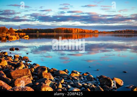 Steine leuchten in der Morgensonne an einem Seestrand. Blauer Himmel mit bunten Wolken, die sich bei Sonnenuntergang in ruhiger Wasseroberfläche spiegeln. Herbstabendlandschaft. Stockfoto