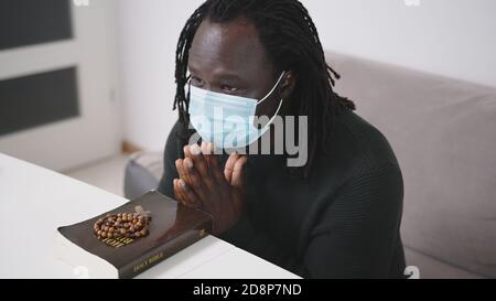 Black African Descent Mann mit Gesichtsmaske Beten auf Heilige Bibel Buch und Rosenkranz. Hoffnung in der Religion während des Ausbruch der Covid-19-Virus-Pandemie. Hochwertige Fotos Stockfoto