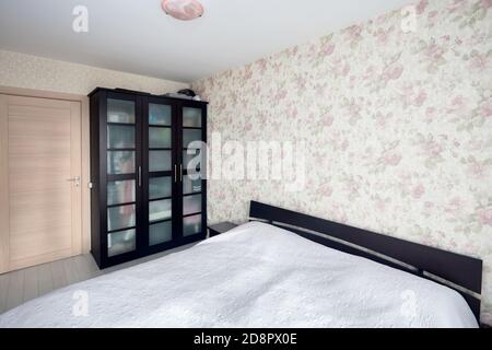 Kleines Schlafzimmer mit einfachem Bett mit Bettüberwurf und geblümten Wänden und Vorhängen Balkon oder Loggia. Konzept der realen Schlafzimmer von Immobilien oder Hotel. Stockfoto