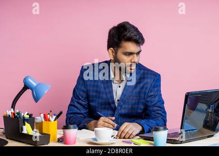 Stilvoller, seriöser junger indischer Geschäftsmann, der am Schreibtisch im Büro sitzt, Karomuster trägt, Smartphone hält, E-Mails überprüft und mobile Ba verwendet Stockfoto