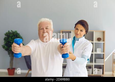 Erfahrene Ärztin unterstützt die Arme des älteren Mannes, während er Hanteln in der medizinischen Praxis hebt. Stockfoto