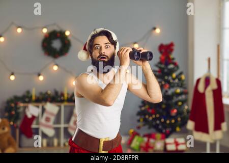 Überrascht bärtigen lustigen Weihnachtsmann in Hause festliche Kostüm stehen Mit Fernglas in den Händen Stockfoto