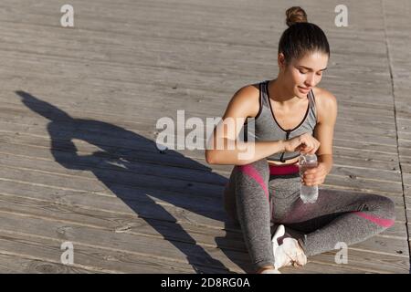 Außenaufnahme einer attraktiven Fitness-Frau, die nach dem Joggen eine Pause macht, auf dem Boden sitzt und Wasser trinkt, am Meer trainiert Stockfoto