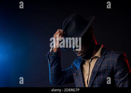 Seitenansicht eines coolen dunkelhäutigen Mannes in stilvollem Anzug, der seinen schwarzen Hut berührt und nach unten blickt. Nahaufnahme des modischen afrikanischen amerikanischen männlichen Modells Stockfoto