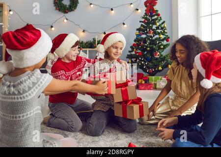 Eine Gruppe von Kindern schenken ihren Freunden am Weihnachtsmorgen Geschenke, die auf dem Boden im Zimmer sitzen. Stockfoto