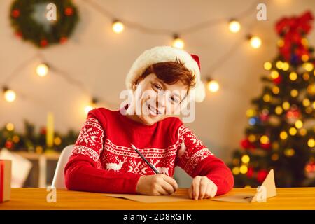 Lächelnder Junge in rotem Hut und Pullover sitzen und schreiben Brief an den Weihnachtsmann mit Wünschen und Träumen Stockfoto