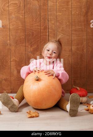 Ein kleines nettes Mädchen in einem rosa Pullover umarmt ein Großer Kürbis, der auf dem Boden auf einem Holzhintergrund sitzt Stockfoto