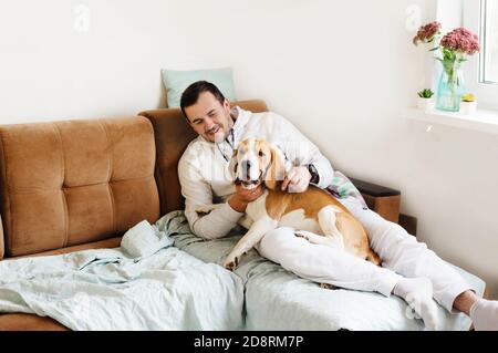 Glücklicher junger Mann mit seinem Hund, Beagle, sitzt auf dem Sofa. Die Jugendart, das Konzept von Haus und Tieren. Stockfoto