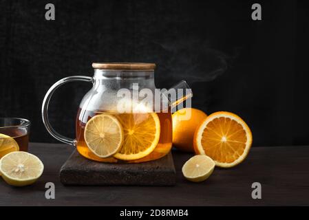 Zitrustee mit Zitrone, Orangen in durchsichtiger Glas Teekanne auf dunklem Holztisch, Kopierraum. Früchtetee. Saisonaler Winter Herbst heißer Drink. Stockfoto