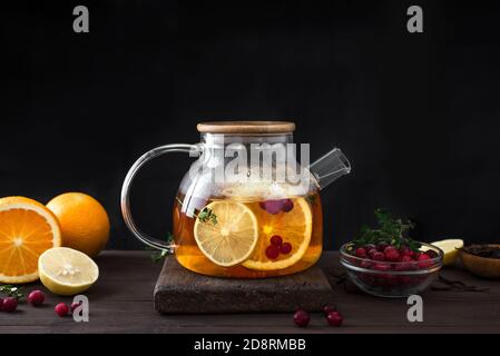 Winter-Früchtetee. Zitrus- und Beerentee mit Preiselbeeren, Zitrone, Orangen in Glas Teekanne auf dunklem Holztisch, Kopierraum. Früchtetee. Saisonaler Winter Stockfoto