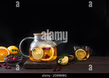 Zitrus- und Beerentee mit Preiselbeeren, Zitrone, Orangen und Thymian in einer Glas-Teekanne auf dunklem Holztisch, Kopierraum. Früchtetee. Saisonaler Winter Herbst Stockfoto